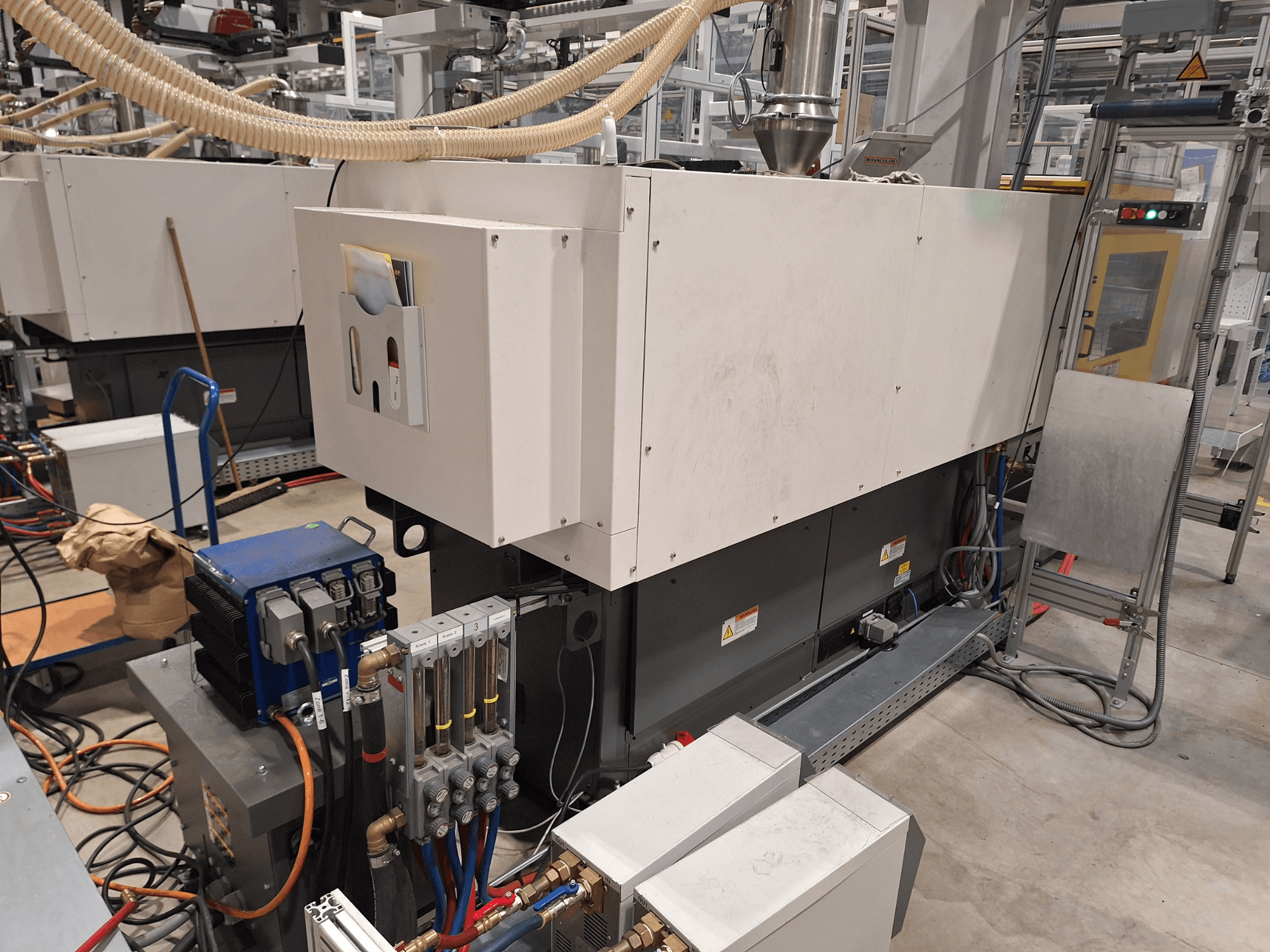 Industriële apparatuur in een fabrieksomgeving; omvat een grote machine met bedieningspaneel, slangen en elektrische aansluitingen, zijaanzicht.