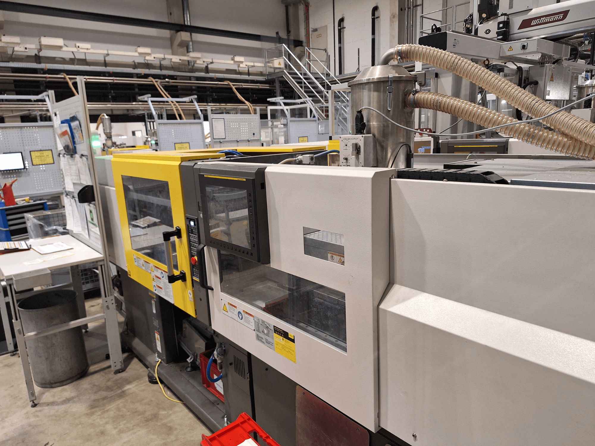 Vooraanzicht van een meerkleurige spuitgietmachine met gele en grijze secties, met bedieningspaneel en uitlaatsysteem.